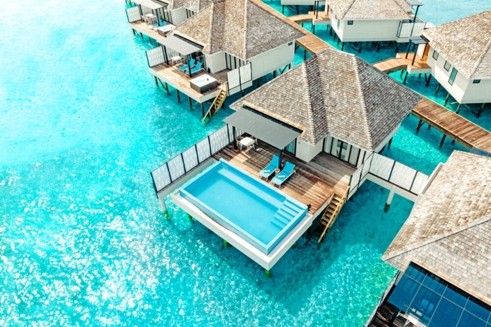 Le ville overwater con tetti di paglia si ergono sull'acqua turchese di questo splendido resort delle Maldive; una villa dispone di un ponte privato con sedie a sdraio e una piscina a sfioro rettangolare con vista sull'oceano.