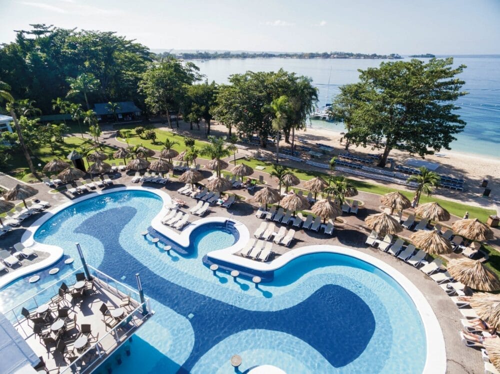 Veduta aerea di un resort con una grande piscina curvilinea circondata da sedie a sdraio e ombrelloni di paglia, situata accanto a una spiaggia sabbiosa fiancheggiata da palme e affacciata sulle calme acque blu dell'oceano.