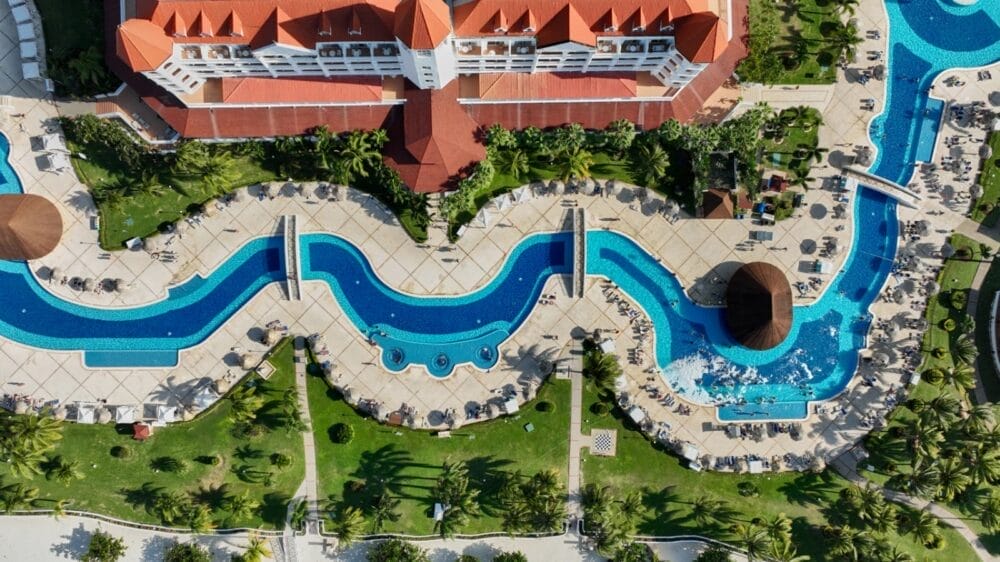Vista aerea di un resort della Giamaica con una grande e sinuosa piscina all'aperto circondata da sedie a sdraio, palme e prati verdi, adiacente all'edificio del Bahia Principe dal tetto rosso.