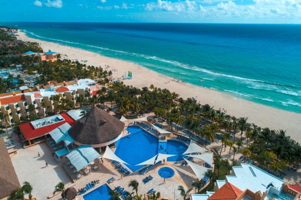 Vista aerea del resort Viva Maya Wyndham con grandi piscine, palme, lettini e spiaggia sabbiosa accanto all'oceano turchese sotto un cielo parzialmente nuvoloso.