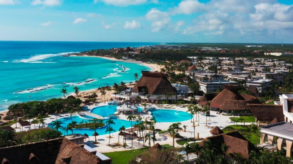 Vista aerea del Bahia Principe Grand Tulum, un resort tropicale con oceano turchese, spiaggia sabbiosa, palme, grandi piscine ed edifici con tetti in paglia, sotto un cielo parzialmente nuvoloso.