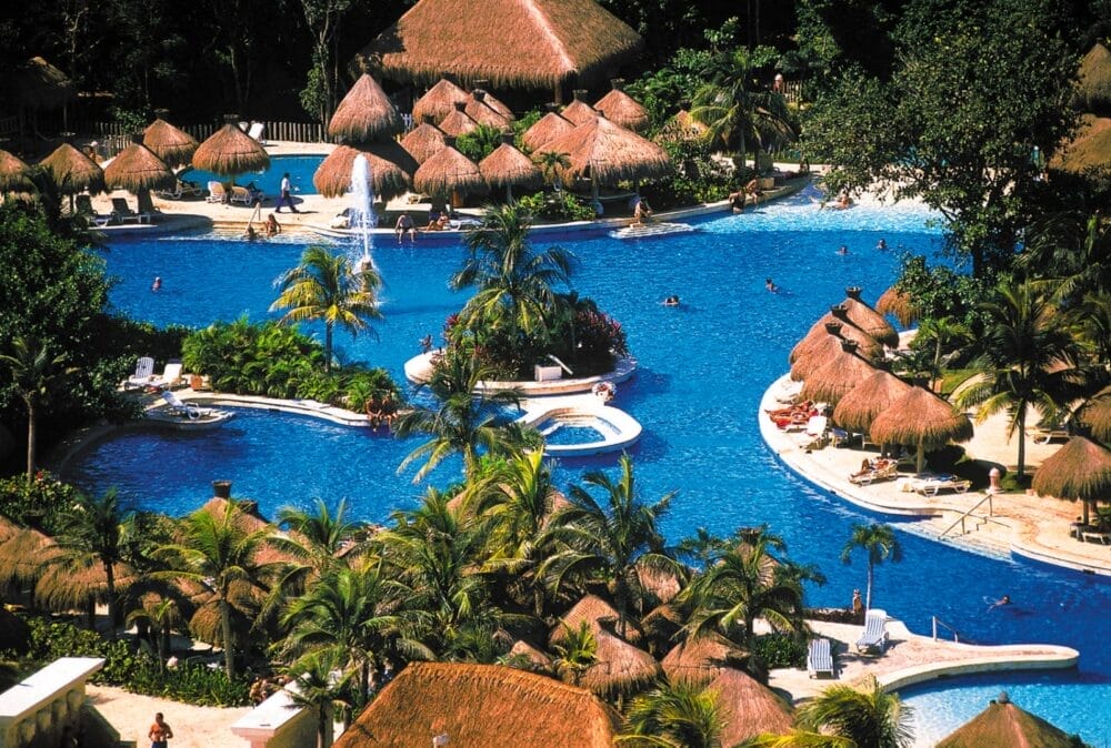 La grande piscina tropicale dell'Iberostar Waves è circondata da palme e cabine con tetto di paglia. Le persone nuotano, si sdraiano sulle sedie e si rilassano al sole. La piscina presenta bordi sinuosi e piccole isole con vegetazione lussureggiante.