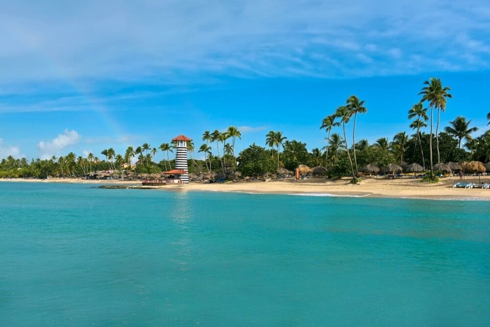 Una spiaggia tropicale con acqua turchese, palme e un litorale sabbioso. Il faro a strisce dell'Iberostar Selection Hacienda Dominicus si erge vicino alla riva sotto un cielo blu brillante con qualche nuvola.