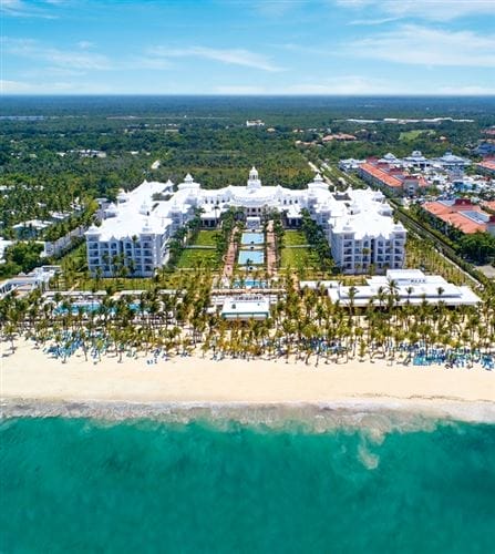 Vista aerea del Riu Palace Punta Cana, un lussuoso hotel all-inclusive con diverse piscine, palme e giardini paesaggistici, situato accanto a una spiaggia sabbiosa e all'oceano turchese, circondato da una vegetazione lussureggiante e da edifici vicini.