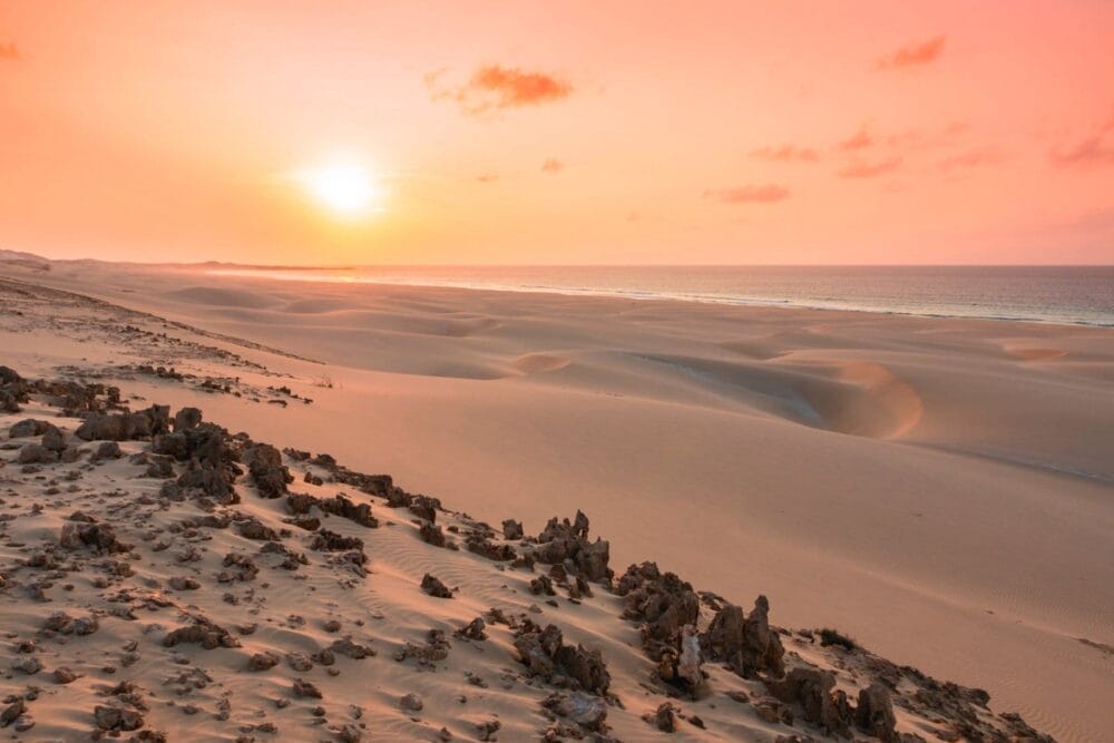 Un vasto paesaggio desertico con dune di sabbia levigata e formazioni rocciose è immerso in una calda luce rosa-arancio al tramonto, che ricorda Bravo Praia De Chaves sotto un cielo parzialmente nuvoloso.