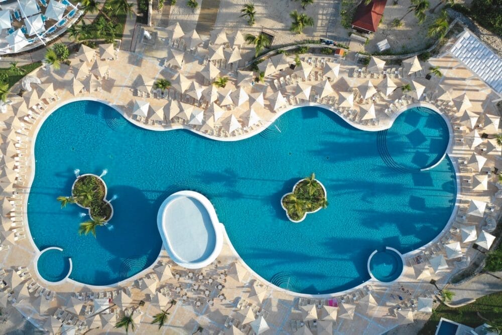Vista aerea della splendida piscina curvilinea del Bahia Principe Luxury Ambar, caratterizzata da due piccole isole di palme, circondate da numerose sedie a sdraio e ombrelloni in una giornata di sole.