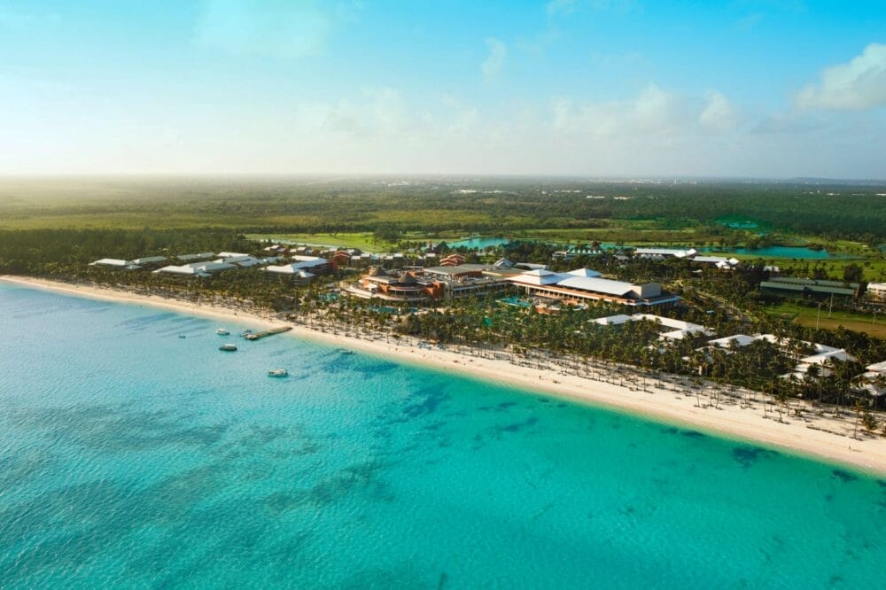 Vista aerea del resort Barceló Bavaro Beach con acqua turchese, spiaggia di Bavaro di sabbia bianca, barche vicino alla riva e un paesaggio verde sullo sfondo sotto un cielo azzurro.