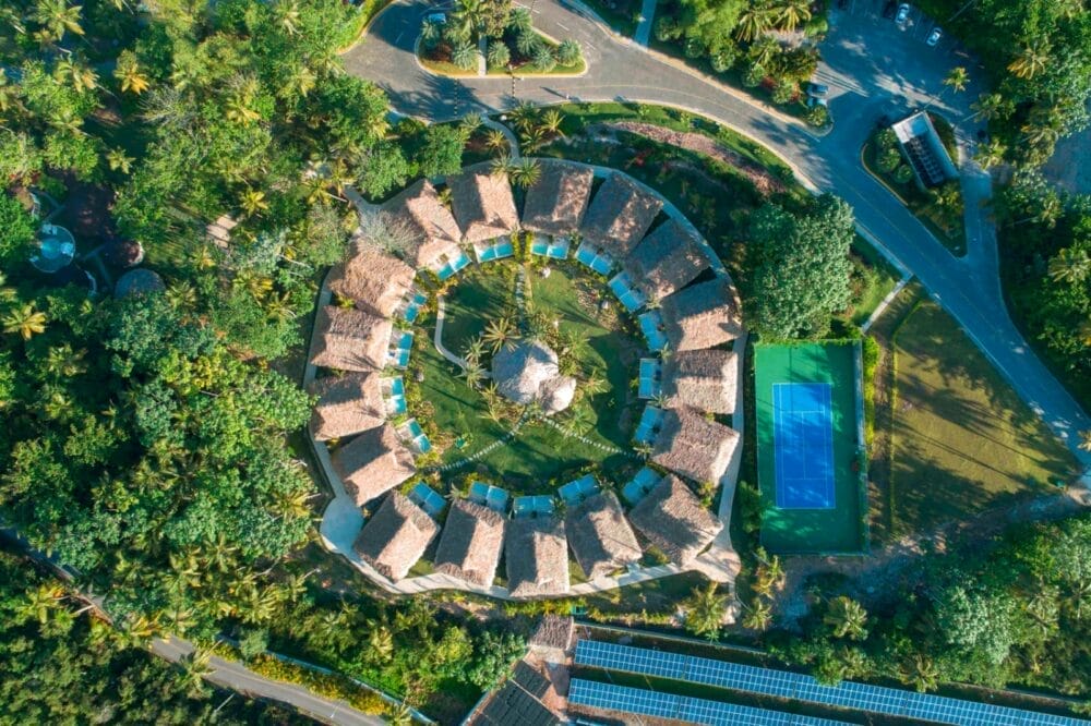 Vista aerea di una disposizione circolare di ville con tetto in paglia al Viva V Samana, circondate da una vegetazione lussureggiante, con un campo da tennis e strade tortuose nelle vicinanze.