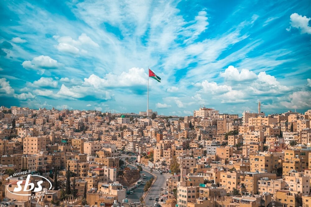 Una vista panoramica di Amman, in Giordania, che mostra edifici beige raggruppati su colline e una grande bandiera giordana, vi invita a iniziare il vostro Tour Magia Del Deserto sotto un cielo blu vibrante con nuvole sparse.