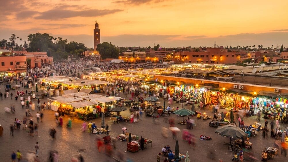 Una vivace piazza del mercato al tramonto con bancarelle luminose, folla sfocata e un'alta torre sullo sfondo, sullo sfondo di un cielo arancione: una scena iconica di un'avventura Viaggio Marocco o Gran Tour Marocco.