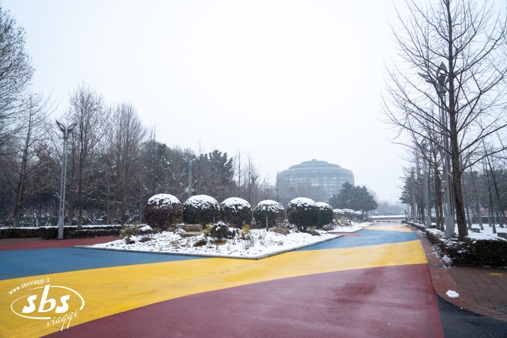 Un ampio sentiero con tratti gialli, rossi e blu attraversa un parco innevato fiancheggiato da alberi e cespugli spogli, evocando i colori di un Tour Cina storica. Un grande edificio a forma di cupola si staglia sullo sfondo, sotto un cielo nuvoloso e invernale.