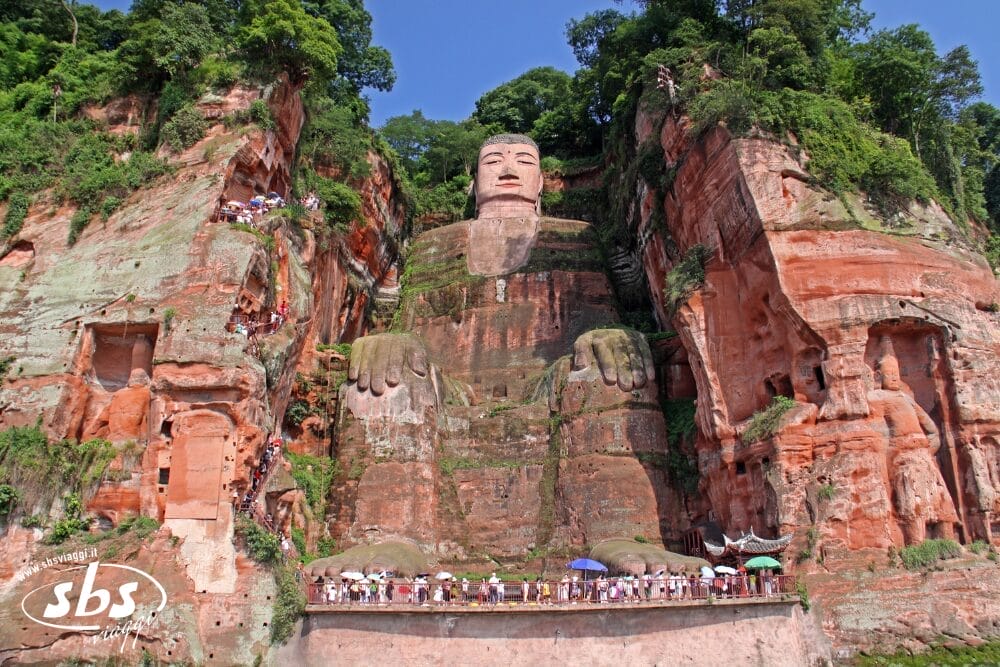L'immagine mostra la gigantesca statua del Buddha di Leshan scolpita in una scogliera rossa nel Sichuan, circondata da una vegetazione lussureggiante, con i turisti dei tour di Tesori del Sichuan in piedi alla base e lungo i sentieri sul fianco della scogliera.