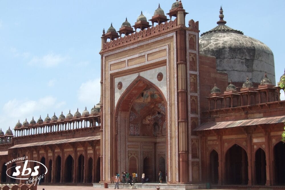 Un grande portale ornato con archi e cupole, costruito in arenaria rossa con accenti di marmo bianco, si staglia contro un cielo blu. Le persone sono visibili vicino all'ingresso, sottolineando la grandezza dell'India Splendida di questa notevole struttura.