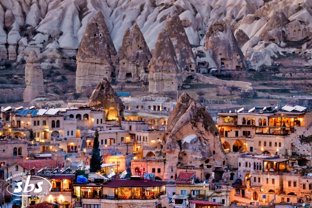 Una vista panoramica della Cappadocia, in Turchia, da un'avventura del Tour Cappadocia, che mostra formazioni rocciose uniche e abitazioni in grotta mescolate con edifici illuminati a giorno al tramonto. Sullo sfondo si ergono alte strutture rocciose appuntite.