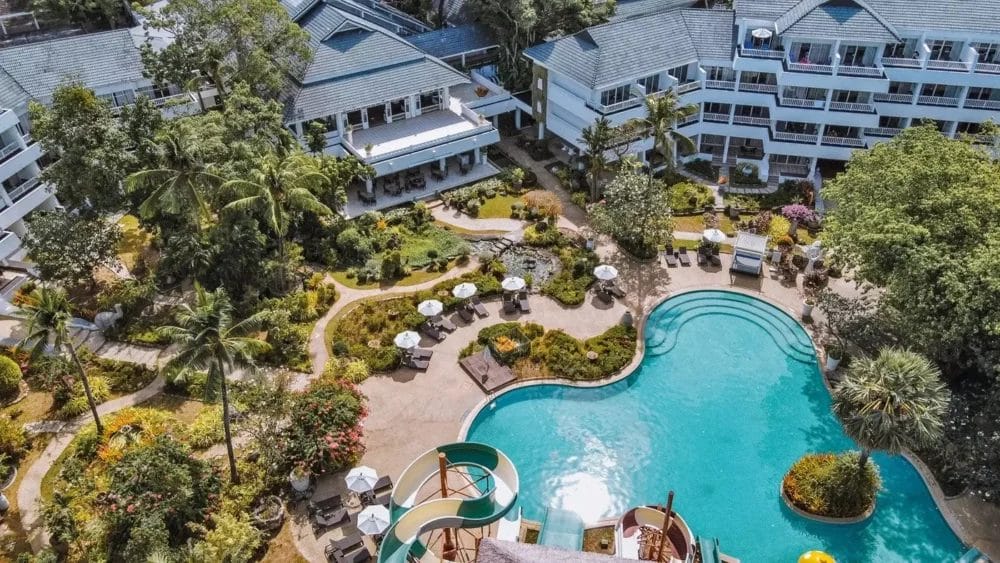 Vista aerea di un resort con edifici bianchi, giardini lussureggianti, una grande piscina all'aperto con scivoli d'acqua, lettini e ombrelloni: la vostra fuga automatica bozza circondata da alberi tropicali.