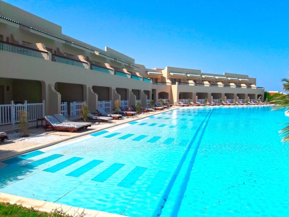 Una grande piscina all'aperto con acqua limpida e blu è fiancheggiata da sedie a sdraio e ombrelloni, accanto al moderno edificio del Coral Sea Holiday Resort, sotto un cielo blu brillante.