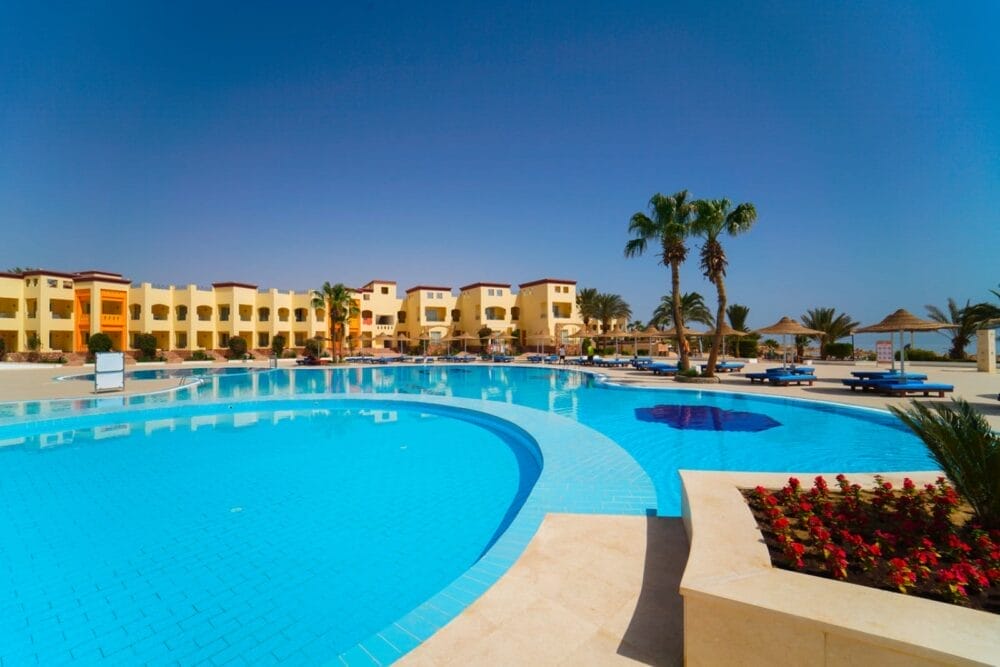 Una grande piscina all'aperto con acqua cristallina, circondata da sedie a sdraio, palme e un'aiuola paesaggistica; gli edifici gialli del resort Ciaoclub e gli ombrelloni di paglia sono visibili sullo sfondo sotto un cielo azzurro e limpido.