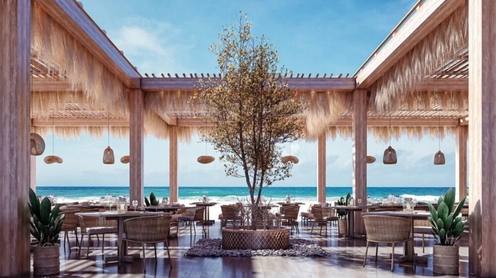 Ristorante all'aperto sulla spiaggia dell'Acel Beach Resort con pergole in legno, sedie intrecciate e arredamento rustico. Un albero centrale è circondato da tavoli da pranzo, con il blu dell'oceano e del cielo a fare da perfetto sfondo all'Alpiclub.
