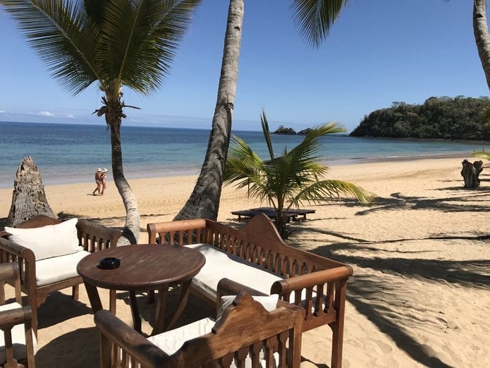 Mobili da salotto in legno per esterni con cuscini bianchi si trovano su una spiaggia sabbiosa sotto le palme del resort Amarina. L'oceano calmo e il cielo blu sono sullo sfondo, con due persone che camminano in riva al mare.