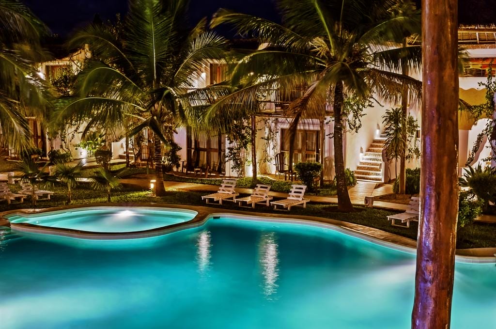 Una piscina serena e illuminata con bordi curvi circondata da palme, sedie a sdraio e una villa a due piani di notte al My Blue Hotel, che crea una rilassante atmosfera tropicale.