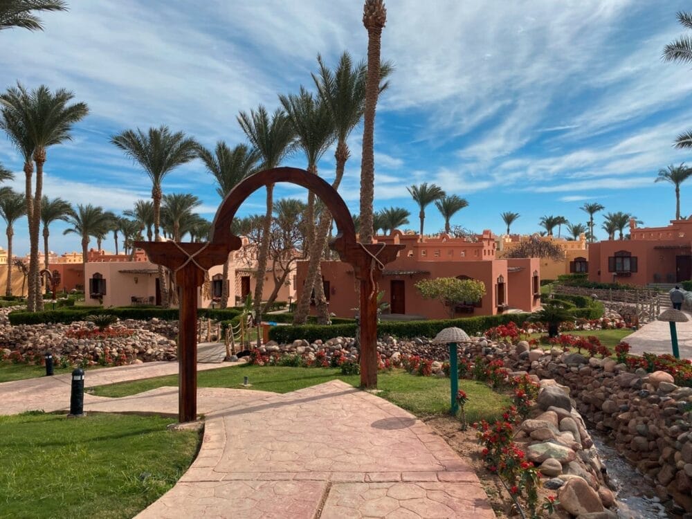 Un sentiero di pietra fiancheggiato da erba e fiori conduce, attraverso un arco di legno, verso gli edifici Bravo Premium Nubian color pesca, circondati da palme sotto un cielo azzurro leggermente velato.