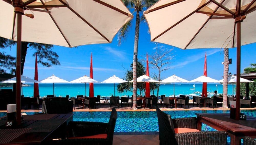 Ristorante all'aperto con tavoli e sedie sotto grandi ombrelloni, affacciato su una piscina e sull'oceano turchese di Kata Beach, con vista su ombrelloni bianchi e barche in lontananza sotto il cielo limpido di Phuket.