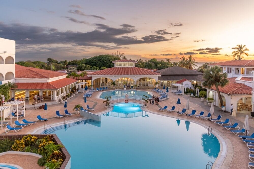 La grande piscina del resort Starfish Cuatro Palmas è circondata da sedie a sdraio blu e palme al tramonto, con edifici dai tetti rossi e un'area bar sullo sfondo: un invitante rifugio Ciaoclub per gli ospiti di età superiore ai 16 anni.