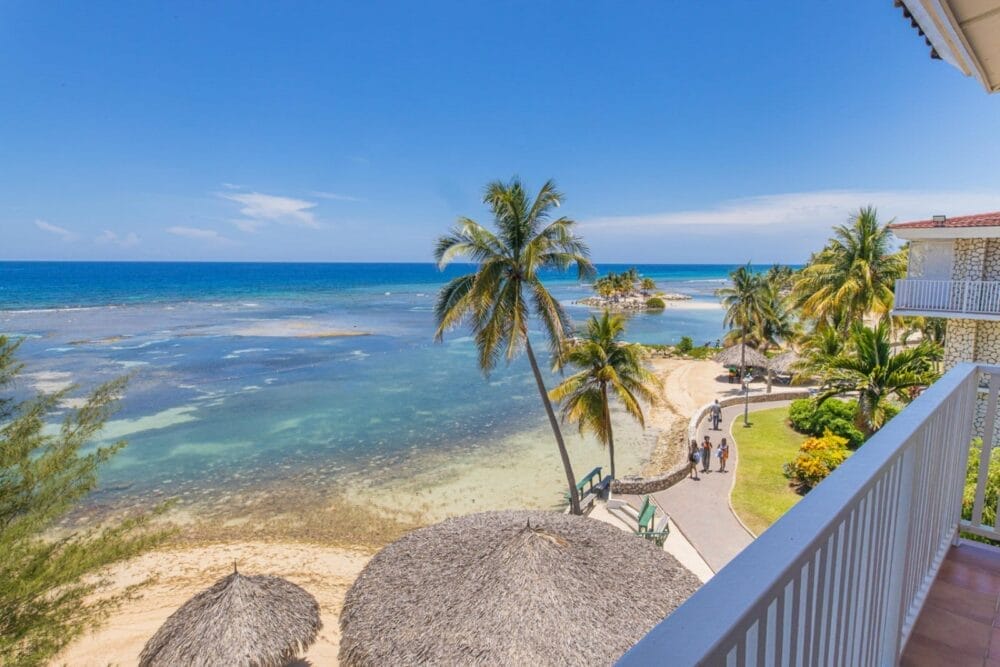 La vista da un balcone del Catalonia Montego Bay si affaccia su una spiaggia tropicale con acqua cristallina, palme, ombrelloni di paglia e alcune persone che passeggiano lungo un sentiero sotto il cielo soleggiato.