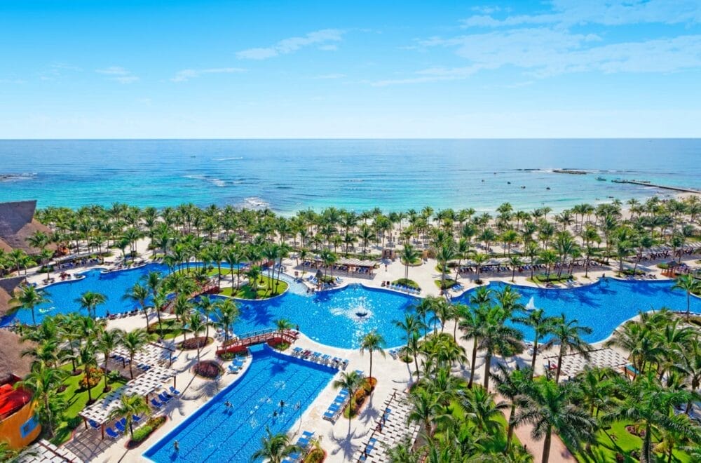 Un grande resort sulla spiaggia come il Barcelo Maya Caribe dispone di diverse piscine circondate da palme, sedie a sdraio e persone che si rilassano, il tutto affacciato su un tranquillo oceano blu sotto un cielo limpido.
