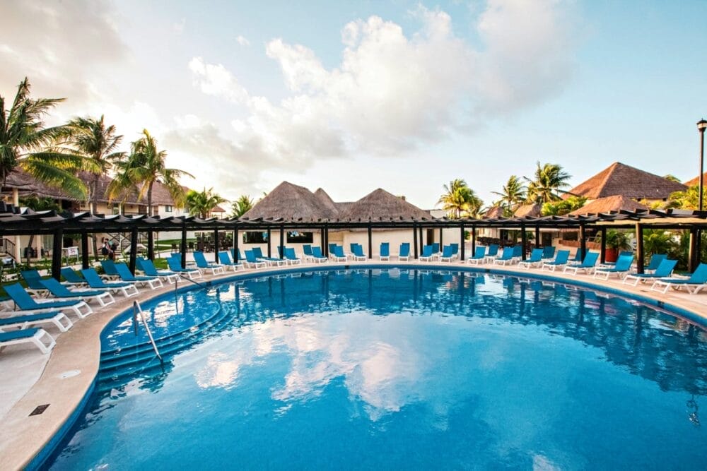 La grande piscina rotonda dell'Allegro Playacar è circondata da sedie a sdraio blu e palme, con edifici dai tetti di paglia sullo sfondo sotto un cielo parzialmente nuvoloso.