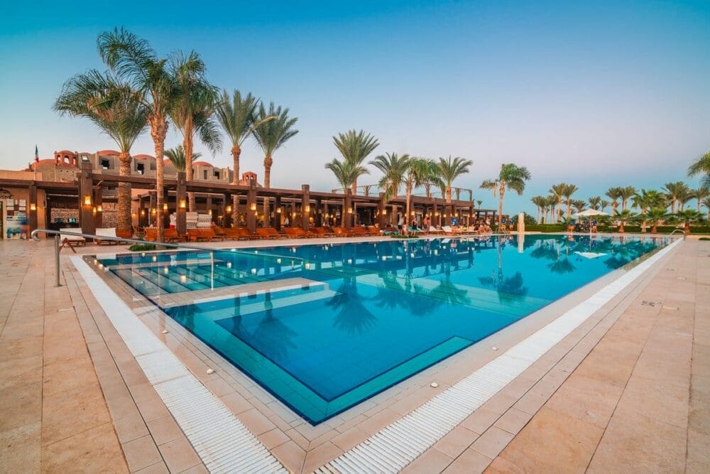 La grande piscina all'aperto del Bravo Gemma Beach Resort è circondata da palme e sedie a sdraio, con l'edificio del resort sullo sfondo, sotto il cielo azzurro del tramonto.