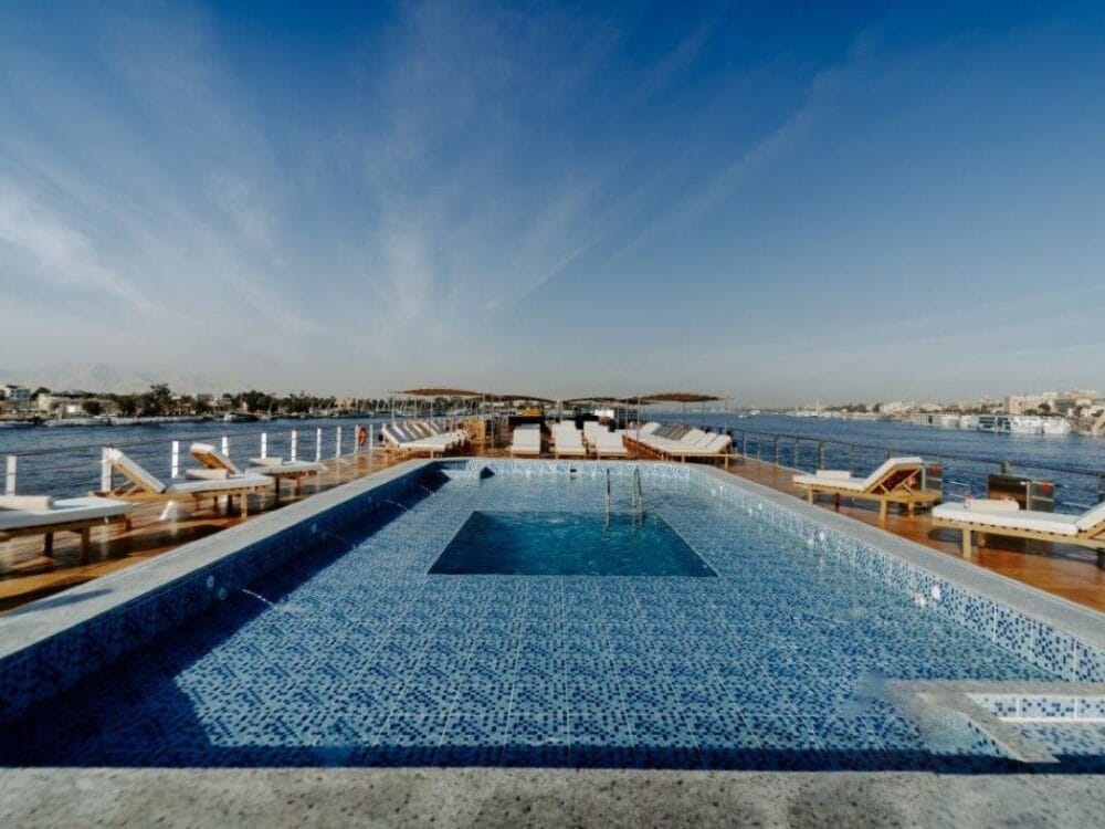 Una piscina sul tetto con piastrelle a mosaico blu si affaccia sul Nilo sulla lussuosa Crociera Nilo. Sedie a sdraio in legno con cuscini bianchi fiancheggiano il ponte, mentre il cielo limpido del Cairo sopra di esso esalta l'atmosfera serena di Le Rive degli Dei.
