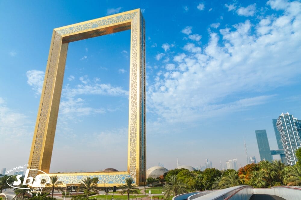Una grande struttura rettangolare dorata, nota come Dubai Frame, si staglia su un cielo blu con nuvole sparse, circondata da palme ed edifici della città: un punto di forza popolare in molte avventure del Minitour Dubai.