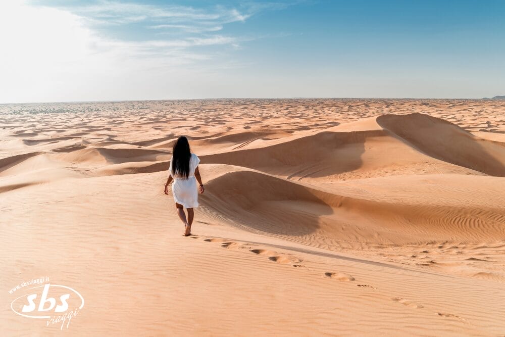 Una donna in abito bianco cammina da sola attraverso dune di sabbia dorata sotto un cielo azzurro e limpido, lasciando dietro di sé impronte nel paesaggio desertico: una delle meraviglie del turismo nel deserto degli Emirati.