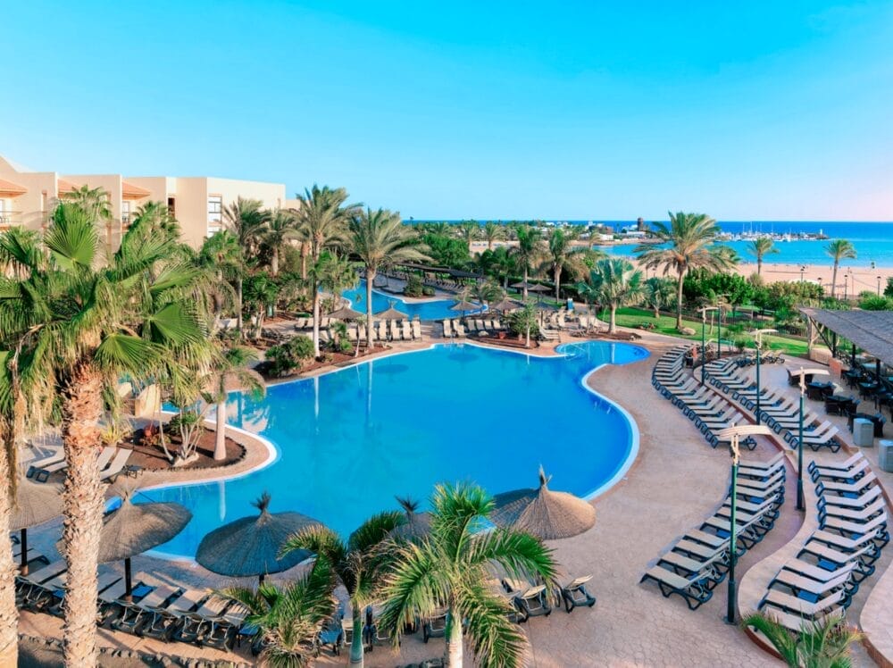 Una grande piscina all'aperto circondata da palme, sedie a sdraio e ombrelloni di paglia al Barcelo Fuerteventura, un resort Alpiclub, con una spiaggia sabbiosa e l'oceano di Fuerteventura visibile sullo sfondo sotto un cielo azzurro e limpido.
