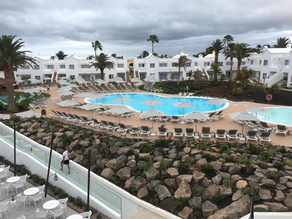 Un resort a Corralejo con edifici bianchi, palme e un'area centrale della piscina fiancheggiata da sedie a sdraio e ombrelloni; paesaggio roccioso in primo piano sotto un cielo nuvoloso.