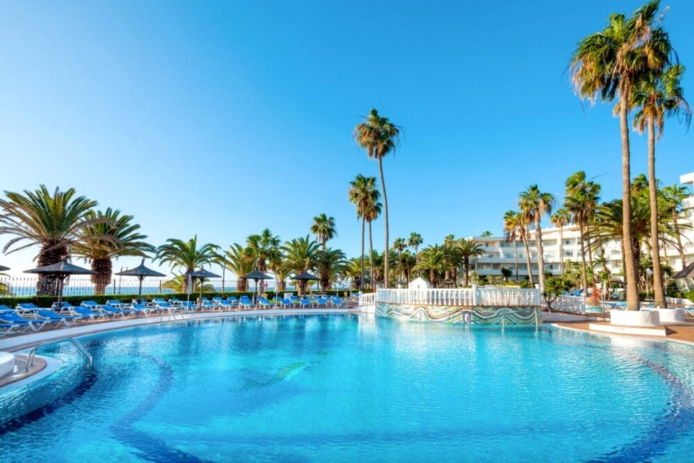 Una grande piscina all'aperto del Sol Lanzarote, circondata da sedie a sdraio blu e alte palme, con un cielo azzurro e l'edificio bianco dell'Alpiclub sullo sfondo.