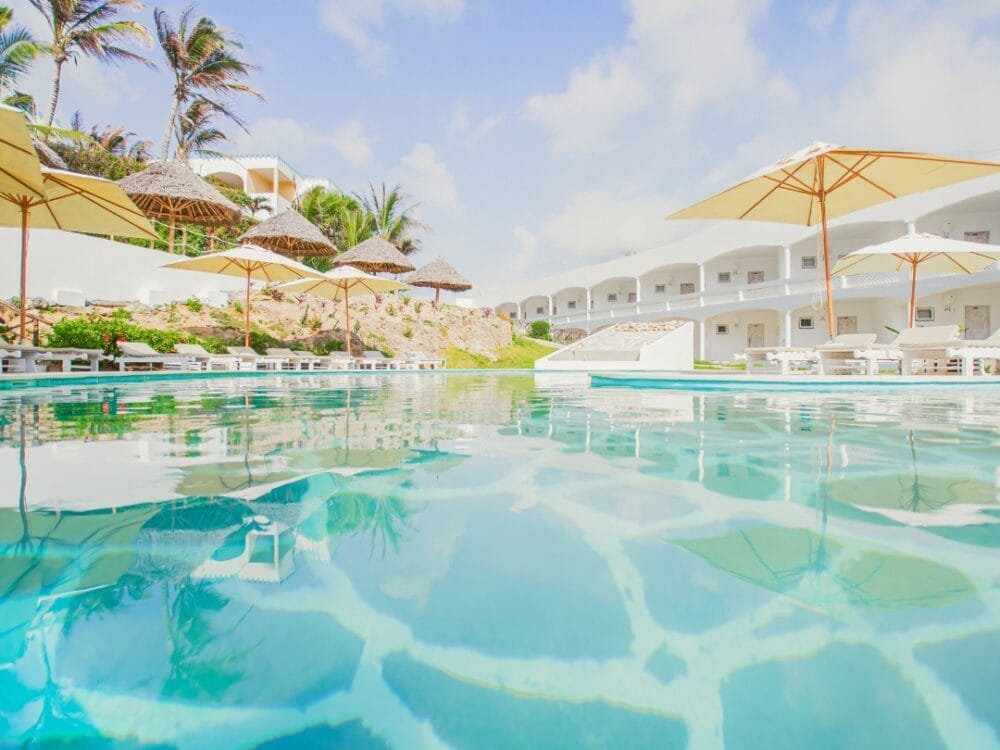 Una piscina trasparente riflette edifici bianchi, ombrelloni di paglia e palme sotto un cielo luminoso e parzialmente nuvoloso al Lion Beach Resort, uno splendido resort 4 stelle ai tropici.