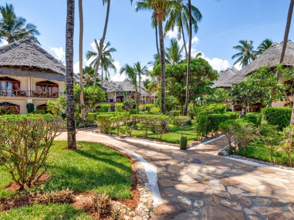 Un sentiero in pietra si snoda attraverso giardini verdi e lussureggianti con cespugli e palme, che conducono a edifici con tetti in paglia sotto un cielo blu brillante al Waridi Beach Resort, un tranquillo resort sulla spiaggia di Zanzibar.