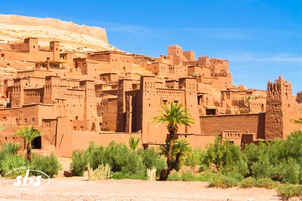 Gli antichi edifici in terra e le torri di Ait Ben Haddou, un villaggio fortificato in Marocco, si stagliano contro un cielo blu brillante con palme verdi: una tappa iconica del Minitour Sahara Express.
