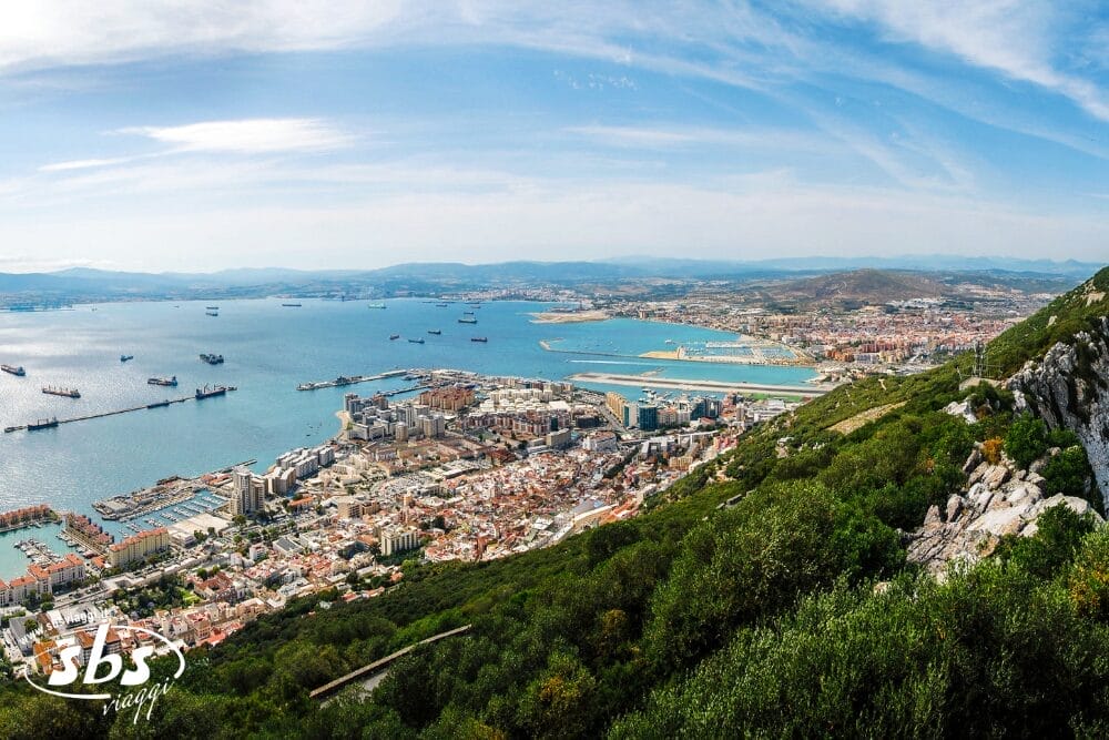 Una vista panoramica di Gibilterra da una collina, che mostra la città, il porto con le navi, il mare blu e le montagne lontane sotto un cielo parzialmente nuvoloso. Una fitta vegetazione ricopre il pendio: una tappa iconica del Gran Tour dell'Andalusia da Malaga.