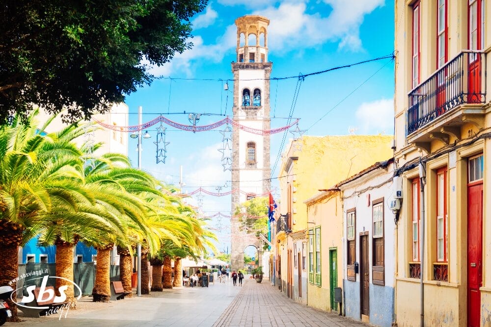 Una strada soleggiata, fiancheggiata da palme e palazzi colorati, conduce a un'alta torre dell'orologio in pietra, ornata di luci e stelle, perfetta per una passeggiata di Meraviglie di Tenerife o di Speciale Capodanno, mentre la gente passeggia tranquillamente lungo il percorso pedonale.