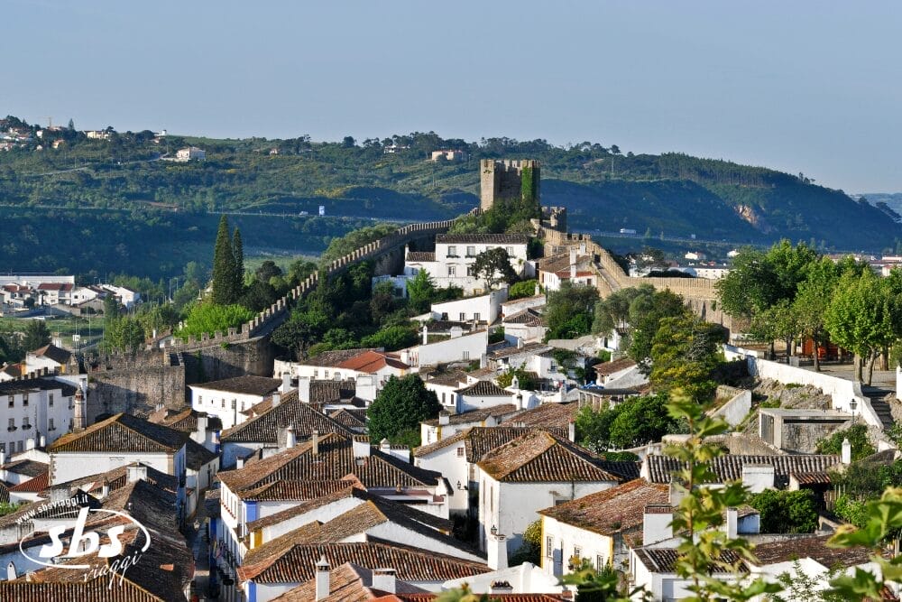 Una vista panoramica di Obidos, Portogallo, che mostra case imbiancate a calce con tetti di tegole e un castello medievale in pietra e mura circondate dal verde - ideale per un minitour o per godersi le feste locali.