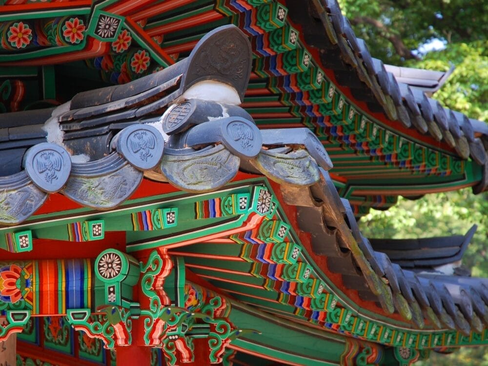 Primo piano della grondaia colorata e ornata di un edificio tradizionale coreano in Corea del Sud, caratterizzato da motivi intricati, vibranti tonalità di rosso, verde, blu e arancione con tegole decorative su uno sfondo verde naturale: un'immagine del sogno coreano.