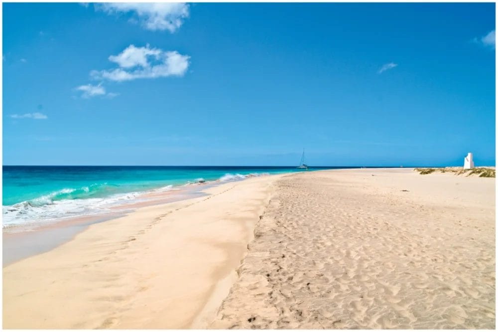Un'ampia spiaggia sabbiosa e vuota a Ponta Sino incontra l'oceano turchese sotto un cielo azzurro e limpido. Onde delicate raggiungono la riva, una barca a vela scivola in lontananza e una piccola struttura bianca si erge sul lato destro della spiaggia.