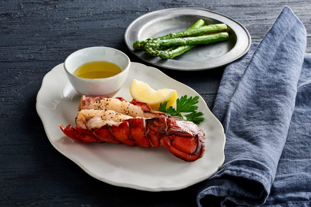 Coda di aragosta cotta con spicchi di limone, prezzemolo e una ciotolina di burro fuso su un piatto bianco. Sullo sfondo, asparagi grigliati serviti su un piatto di metallo accanto a un tovagliolo blu, perfetto per una cena automatica Bozza.