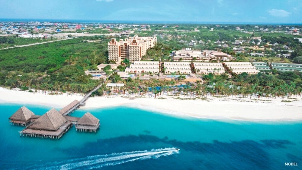 Veduta aerea del resort sulla spiaggia del Riu Jambo Hotel con capanne dai tetti di paglia sull'acqua turchese, un lungo molo, una spiaggia di sabbia bianca, una vegetazione lussureggiante e un paesaggio urbano sullo sfondo sotto un cielo azzurro e limpido.