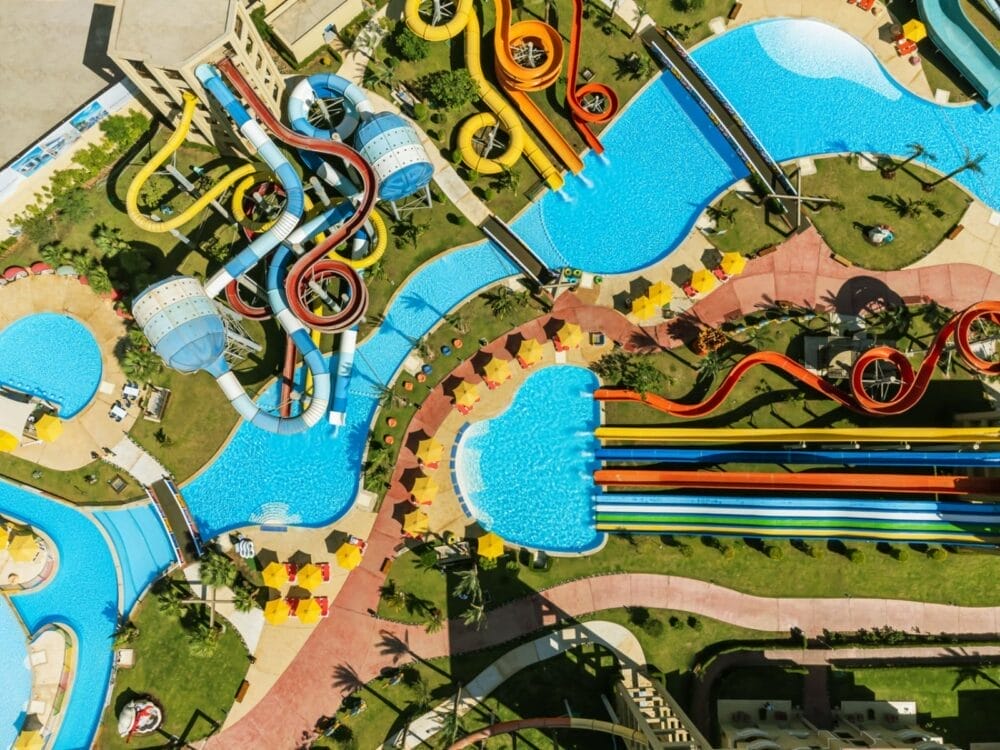 Vista aerea del coloratissimo parco acquatico Rixos Premium Seagate, con scivoli sinuosi, lettini, ombrelloni e grandi piscine blu circondate da erba verde e palme.