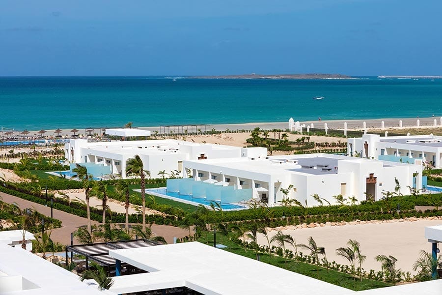 Ville bianche e moderne sulla spiaggia, con piscine private e palme, si affacciano su una spiaggia sabbiosa e sull'oceano turchese, sotto un cielo azzurro e limpido, nell'elegante Riu Palace Boavista, un resort di Boavista che offre lusso da hotel all-inclusive e viste mozzafiato sull'isola.