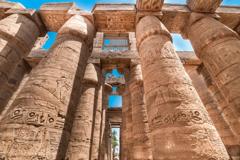 Le massicce colonne di pietra antiche, ricoperte da intricati geroglifici egiziani, si innalzano verso il cielo di Luxor sotto la luce del sole, con il cielo blu e le palme sullo sfondo, a ricordare la grandezza dell'epoca di Ramses.